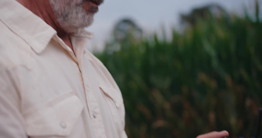An older male is holding and operating a drone controller in his hands he is standing next to a cornfield