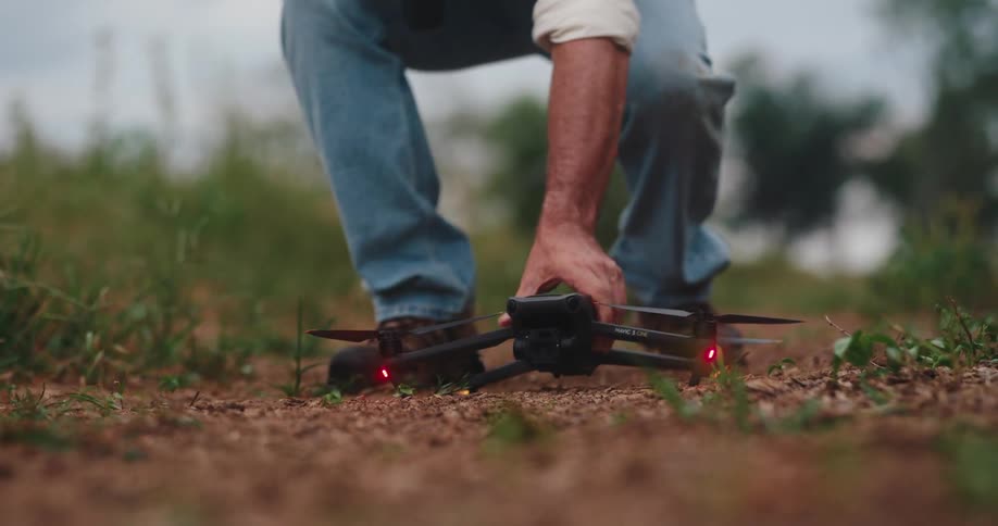 The video shows a lowangle shot of a person wearing jeans and work boots walking on a dirt path surrounded by grass the person stops bends down and places a black drone on the groundthe drones lights are illuminated suggesting it is being prepar
