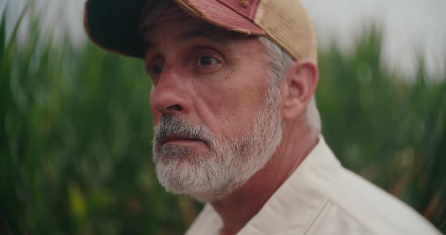 Older farmer standing near a field of crops glances up with a thoughtful expression
