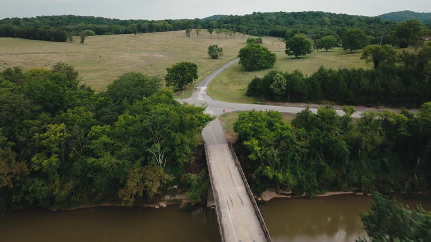 Narrow bridge crosses a green area with a winding road in the background