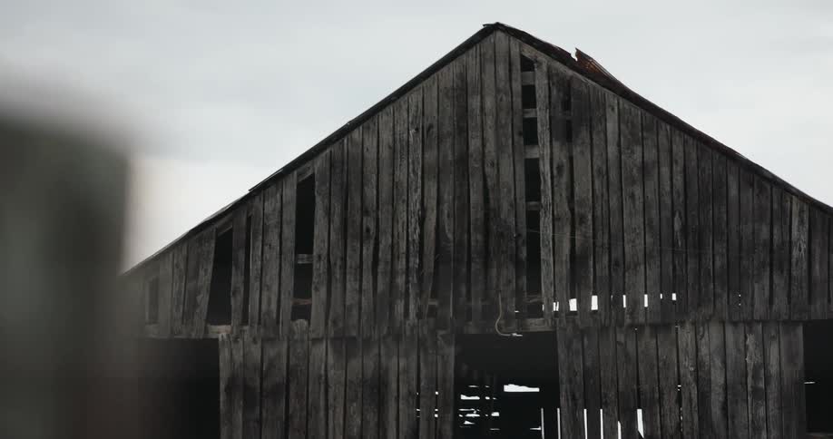 The video features an upward panning shot revealing the weathered dark wooden facade and triangular gable roof of an old barn or agricultural structurethe structure shows signs of age with missing or separated planks allowing slivers of light and gli