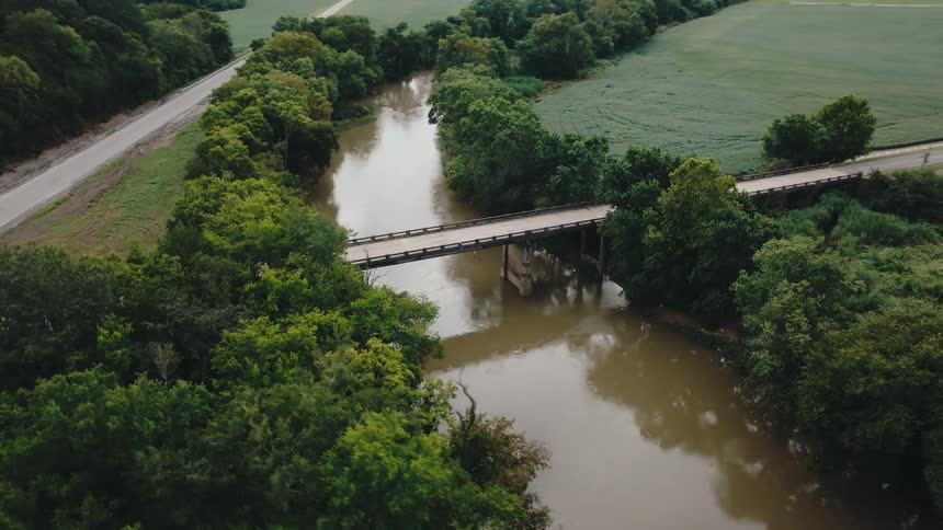 A wooden bridge spanning a muddy river surrounded by dense green trees and bordered by lush green fields and a narrow road