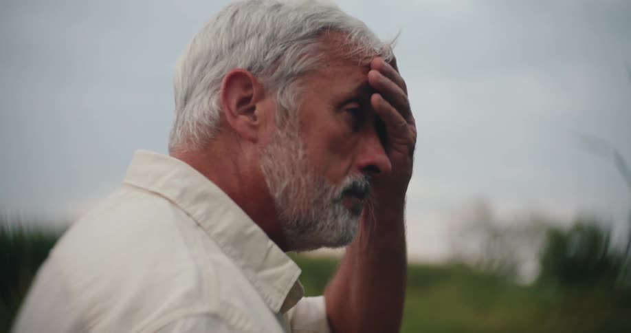A greyhaired farmer removes his baseball cap rubbing his head and face with a tired or thoughtful look before putting the cap back on to gaze over his field