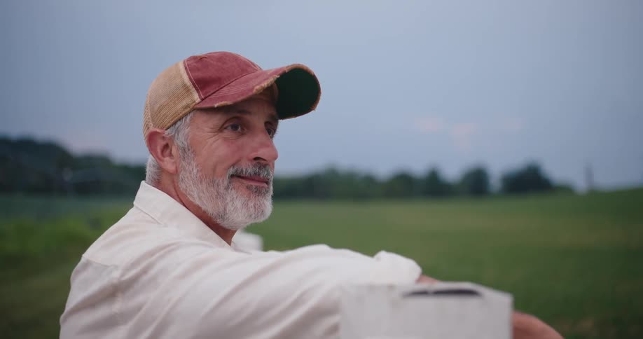 A smiling man leans on a fence overlooking a grassy field at dusk