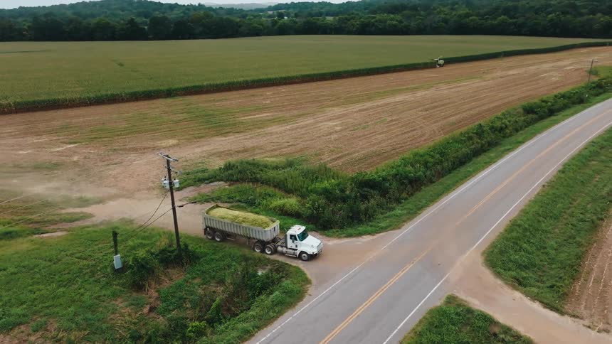 Aerial shot of a dump truck full of crops driving on a rural road beside a large farm field