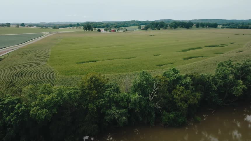 An expansive view of rolling green farmland reveals a river in the foreground and a distant red barn