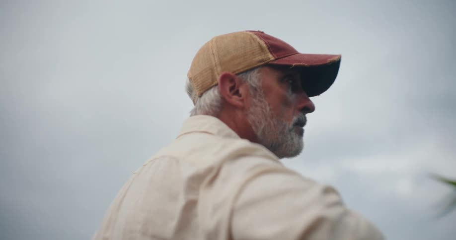 Older farmer standing near a crop field wipes his sweat before walking away on an overcast sky