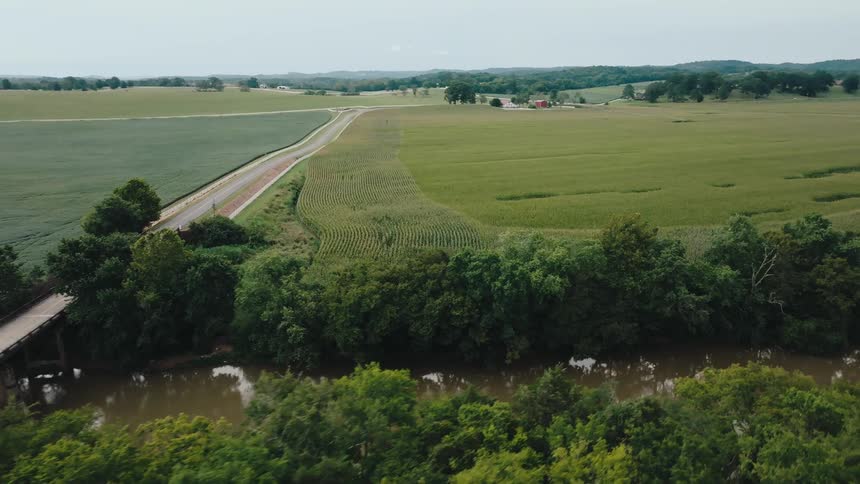 A road crossing a narrow bridge over a dark winding river bordered by thick trees and an expansive rural landscape dominated by large geometrically shaped fields of green crops and rolling distant hills under a diffused bright sky