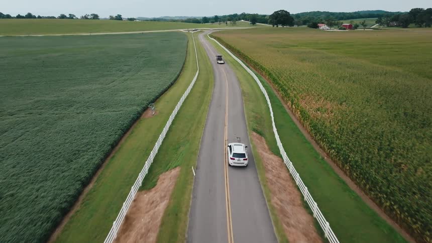Cars drive along a rural road bordered by fields and white fences