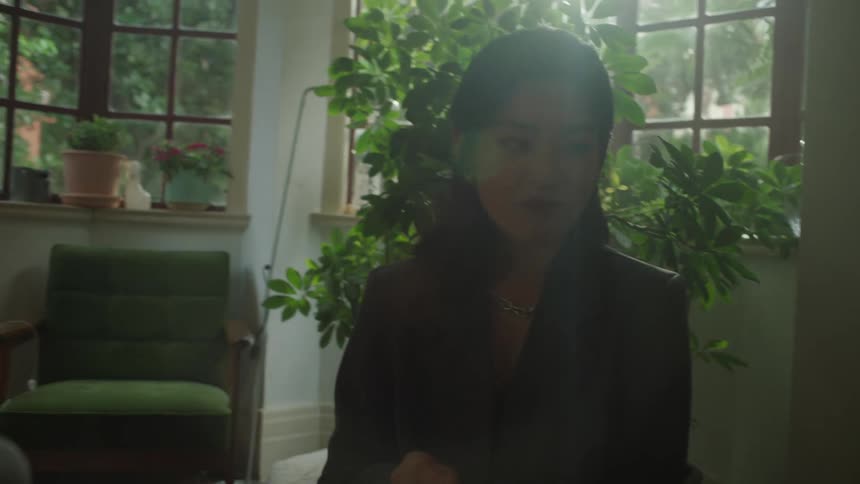 A woman in a dark blazer with a subtle silver necklace sits in a dimly lit bohemian room surrounded by indoor plants as she speaks and holds a small white object during a conversation with an unseen person