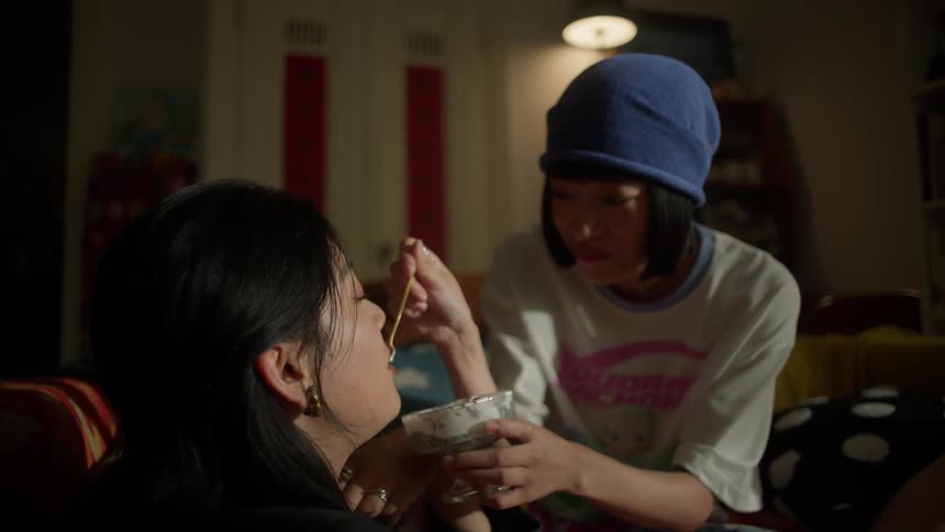 A person in a blue beanie gently feeds another person from a bowl under warm cozy room lighting creating an intimate and caring atmosphere