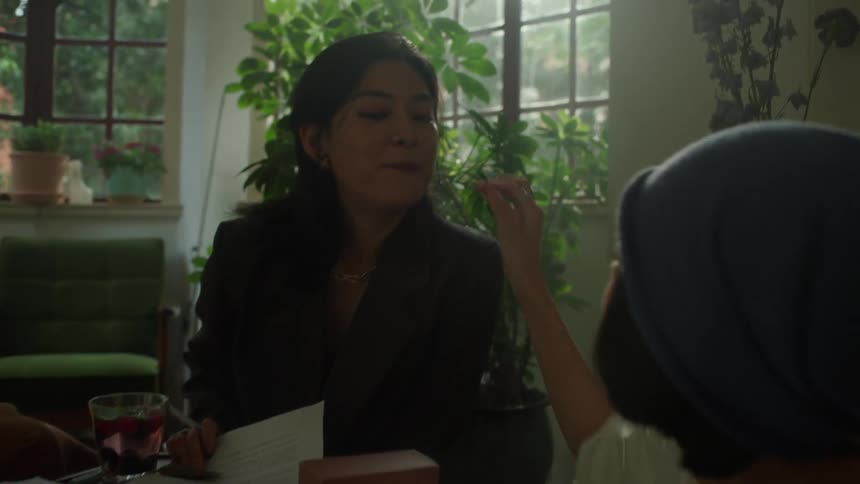 A woman sitting at a table in a sunlit room smiles and talks while chewing after being fed a bite of food by a friend sitting across from her