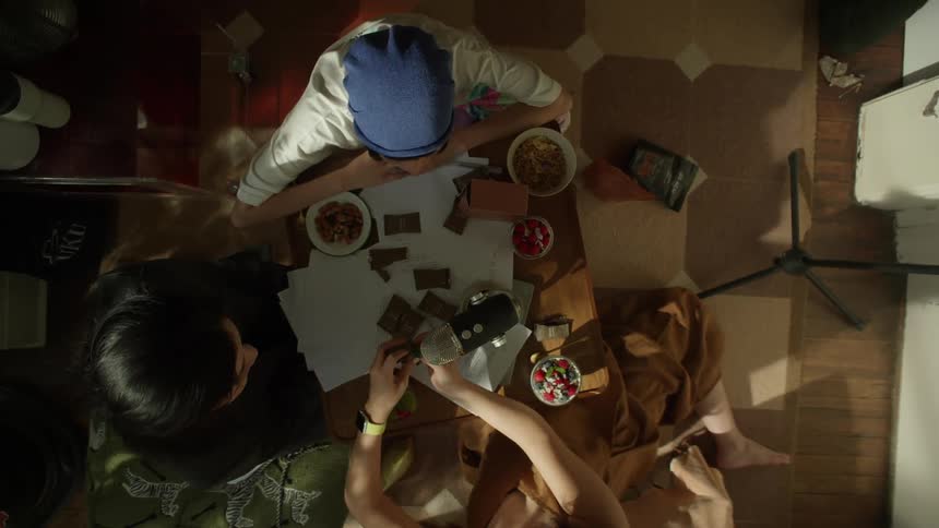 An overhead view shows three people gathered around a small table covered with papers bowls of food and colorful candy recording sounds in a warmly lit indoor setting