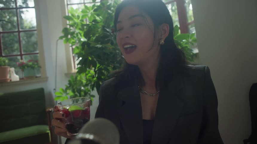 A young asian woman wearing a suit sits indoors at midday enjoying a drink while speaking into a microphone during a podcast