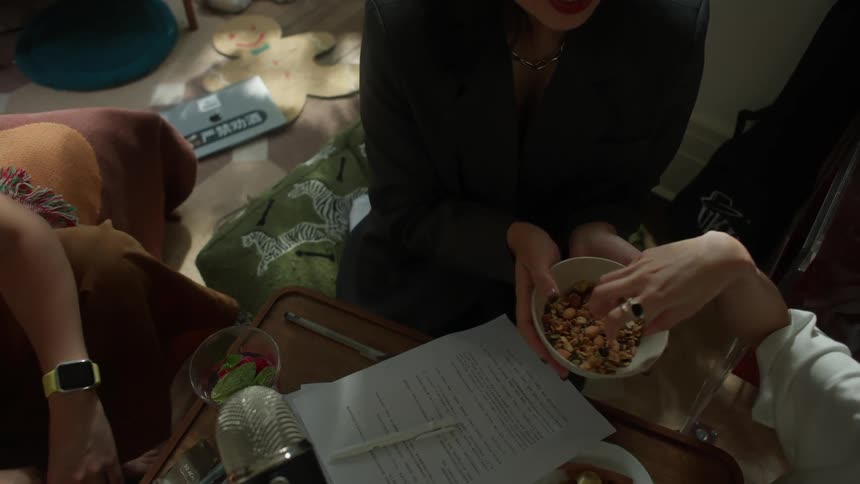A close view shows three people sitting around papers and food with one woman holding a bowl of granola recording its sounds on a microphone visible on the table