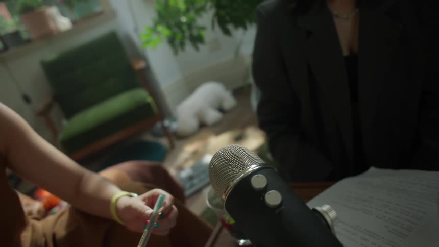 A small table where hands belonging to several individuals one wearing a dark blazer and holding a pen and another holding a clear glass bowl of berries are clustered near a large professional recording microphone