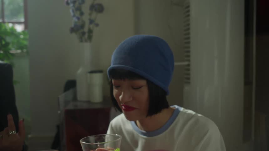 Three friends share a comfortable moment indoors as a woman in a black blazer takes a drink and another woman in a blue beanie sips from a glass filled with berries near a bright window