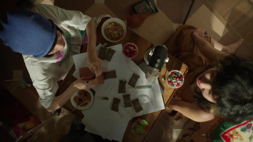 Three people eating sit around a small table scattered with papers and wrapped chocolate bars surrounded by bowls of snacks and a central microphone
