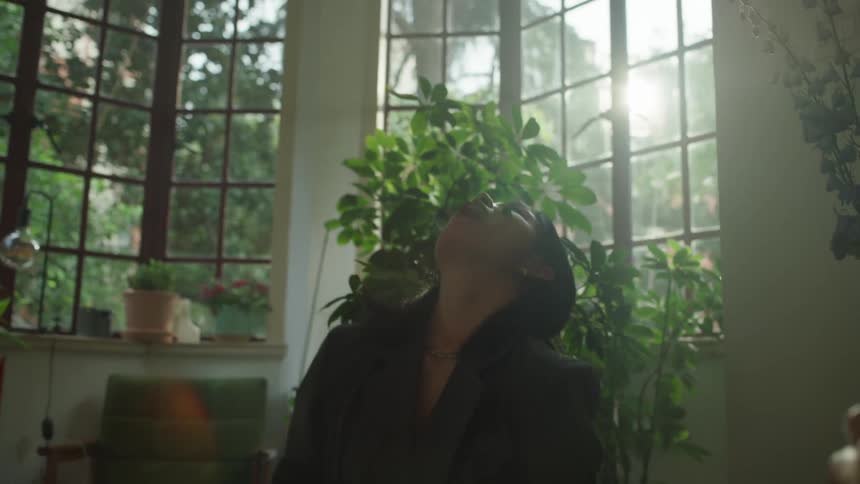 Two women sit in a sunlit room filled with lush plants laughing as one female tries to throw food into her mouth