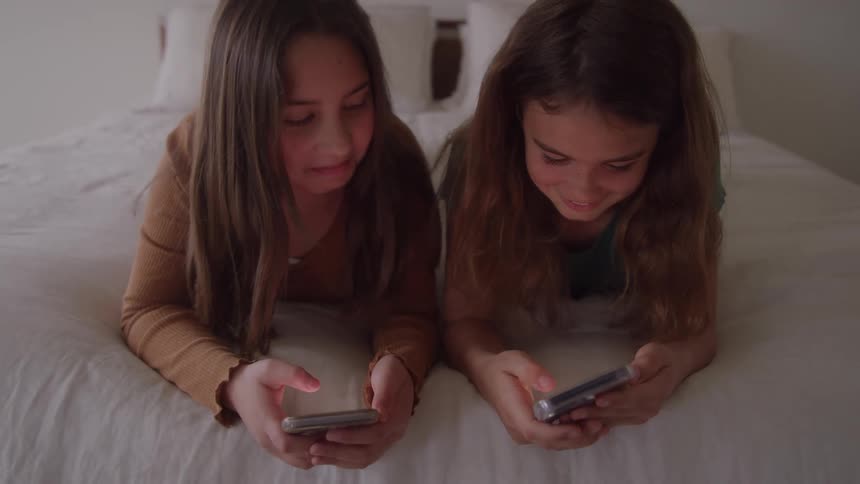 Two young girls lie sidebyside on a white bed smiling as they look down and interact with their smartphones in the soft light