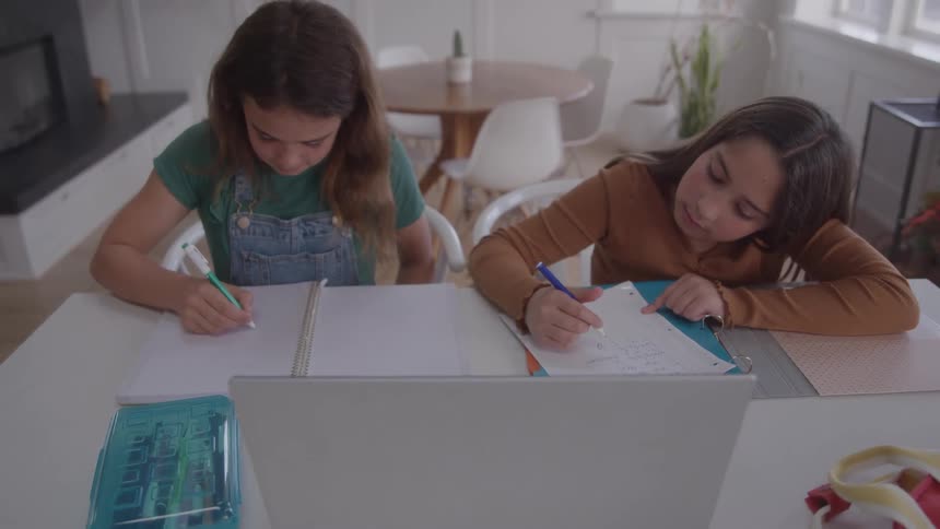 Two little girls are studying while writing in notebooks in front of a laptop