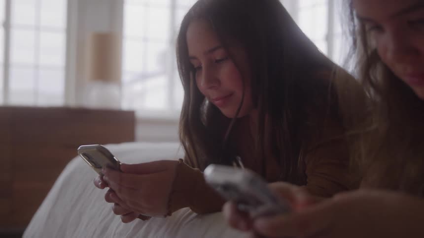 Two young women lie side by side on a bed both focused on their smartphones
