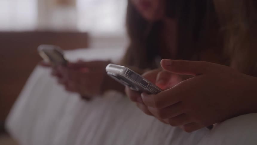 Two girls lying on a bed and using their smartphones have a conversation