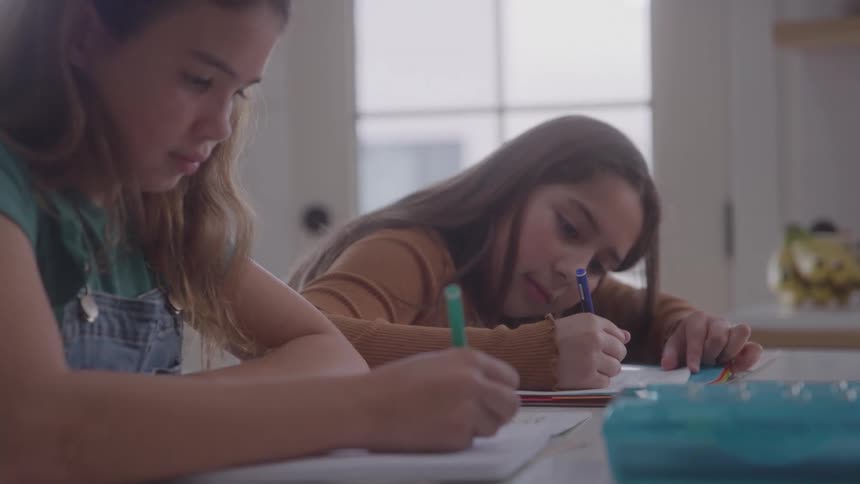Two young girls are in a kitchen sitting at a table coloring in books