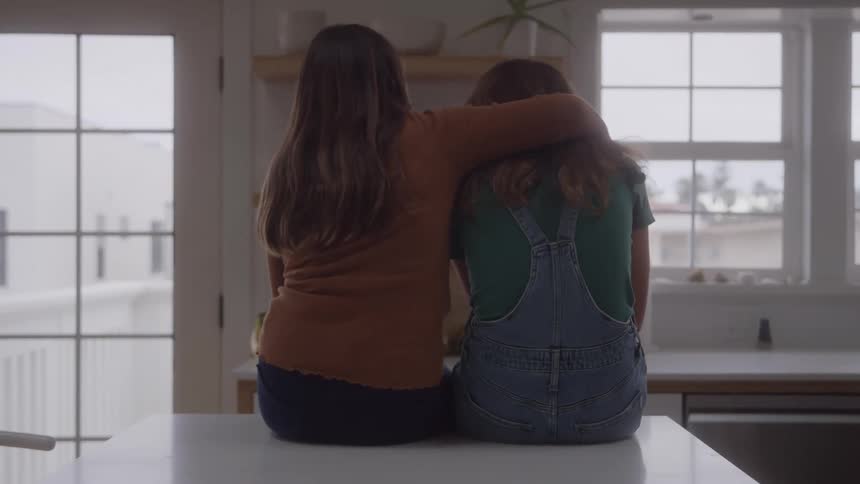 Two teenage girls sitting together on the kitchen counter and one girl puts her arm around the sad girl to comfort her