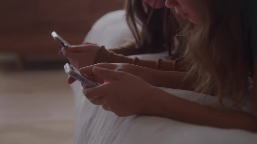 Two girls lying on the bed use their phones side by side