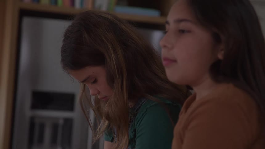 Two girls rest their heads together while one appears focused on what she is doing