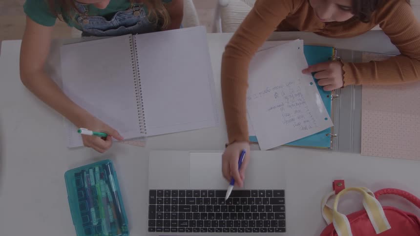 Two girls sit together at a table referencing a computer while they work on their homework