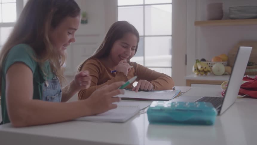 Two young girls sitting at a table with a laptop computer writing in notebooks and laughing