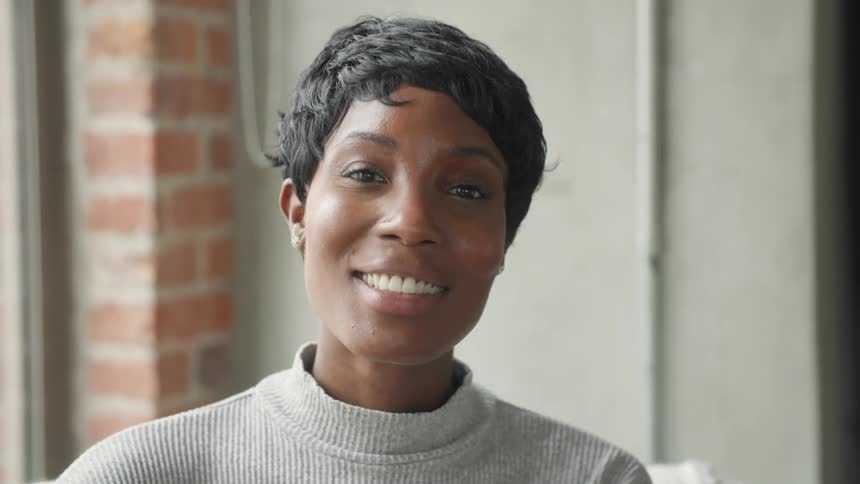 A woman with short hair and a grey sweater smiles warmly while looking directly into the camera her face lit by soft indoor light from a nearby window