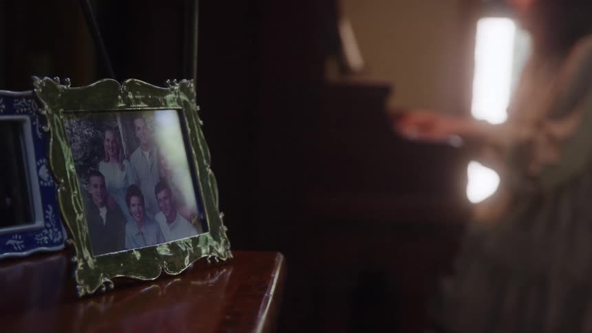 A family photo in a gold frame sits on a table with a person in the background playing the piano