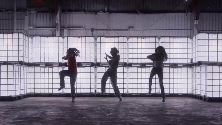 Three dancers rehearse choreography in a warehouse lit by large light panels