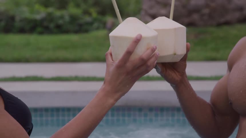 The side of a male and females body are shown toasting coconut drinks while in a pool