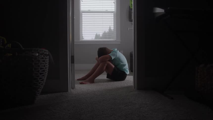 Boy sitting on the carpeted floor hides his face in the arms resting on his knees during the day