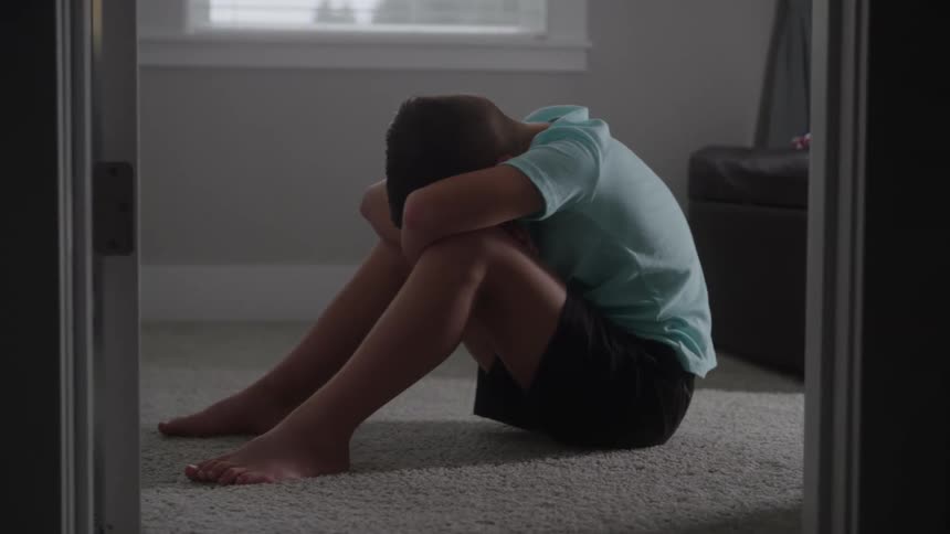 A sad young boy sits alone on the floor of his room and buries his head in his arms