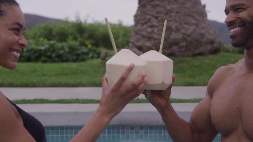 A man and woman in a pool clink coconuts together smiling happily at each other with palm trees and distant mountains in the background