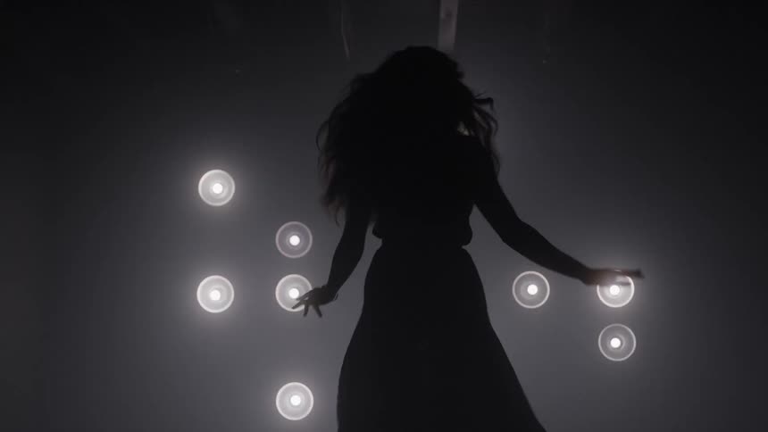 A womans silhouette moves gracefully in deep darkness passing by several bright round lights before disappearing from the frame