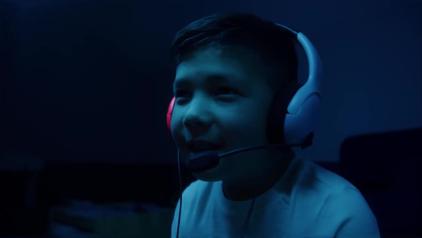 A young boy intensely focused on a screen outside the frame wearing a large white gaming headset with a boom microphone his face and white shirt brightly illuminated by a harsh dramatic neon blue light as he speaks into the microphone