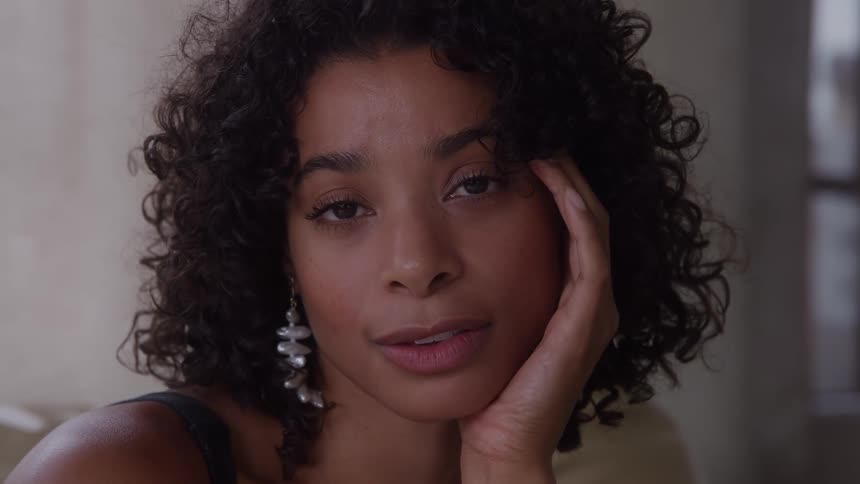 A young black woman sitting on a chair indoor with long curely hair wearing long earrings midday