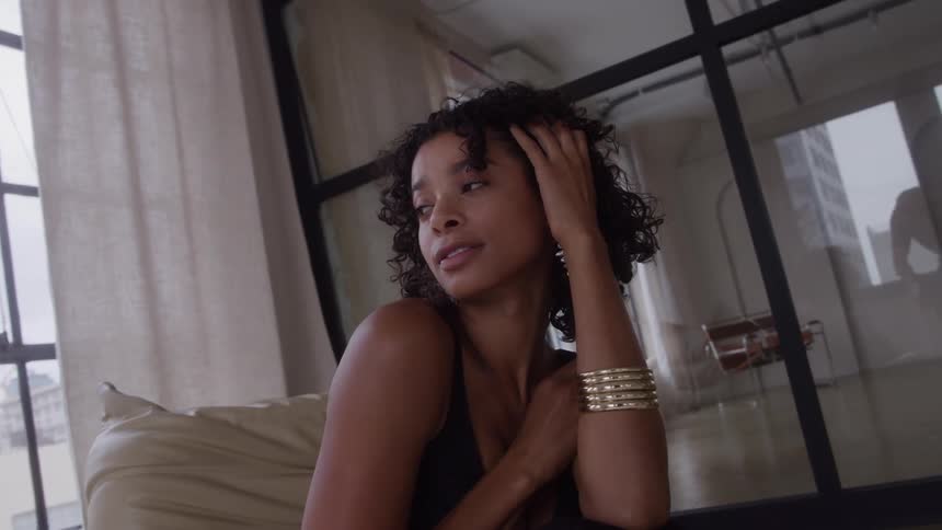 In a modern apartment setting a poised woman with an elegant black dress and gold bracelets sits in a chair and gently touches her curly hair