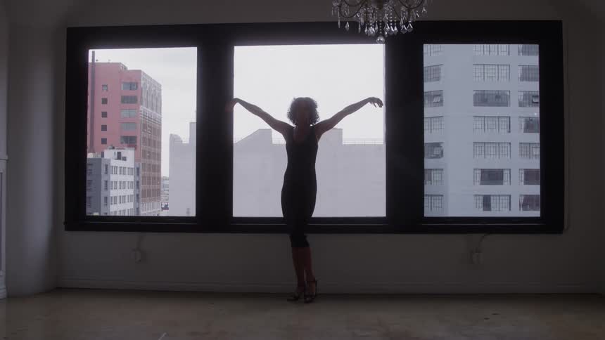 Woman poses with arms open in front of large windows overlooking the city
