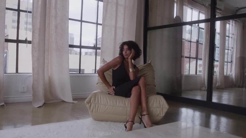 Woman sits on a beige chair posing in a studio filled with large window light