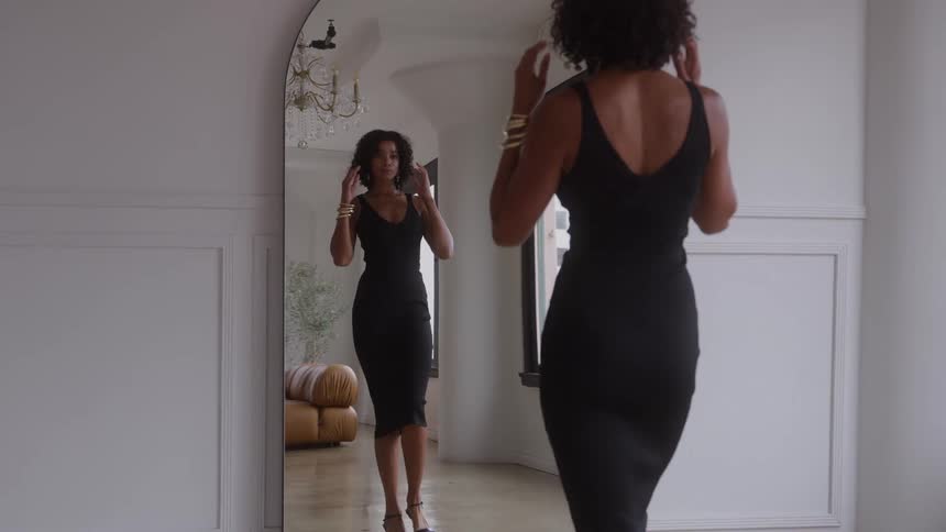 A woman in a black dress looks at her reflection in a full length mirror and adjusts her hair