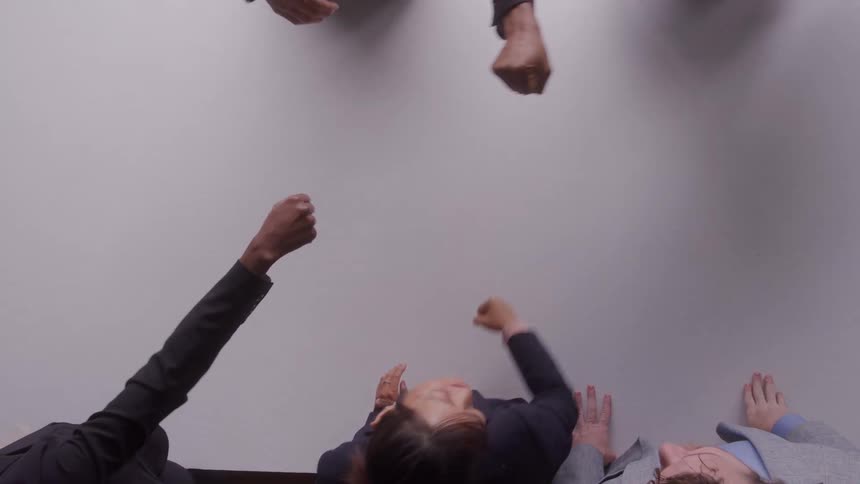 Seen from overhead five people reach their fists toward the center of a white table bumping them together in a group gesture of teamwork