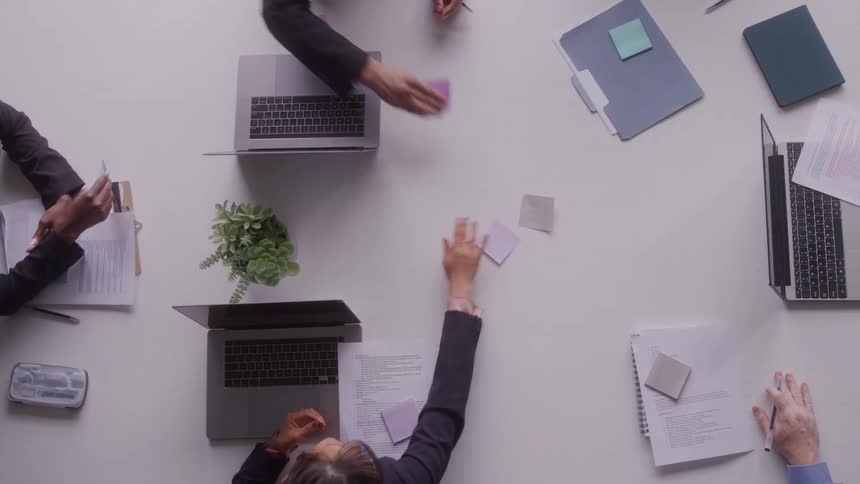 An overhead view shows several people seated around a white table covered with laptops documents folders sticky notes and a small potted plant as they reach toward the center to stick post its on the table