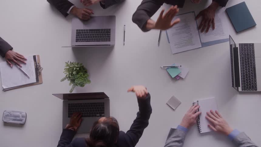 A topdown view captures a busy meeting as colleagues gesture type on laptops and review documents spread across a white conference table